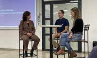Drei Personen sitzen an einem hohen Tisch und sind im Gespräch. Im Hintergrund läuft eine Powerpoint-Präsentation.