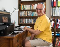 [Bitte in "Französich" uebersetzen:] Ein Mann sitzt vor dem Computer und lächelt in die Kamera