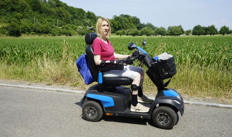 Betroffene einer Hirnverletzung Eine Frau mit blonden Haaren sitzt in einem Elektro-Rollstuhl vor einem dicht bewachsenen Feld. Ihr Gesichtsausdruck ist ernst.