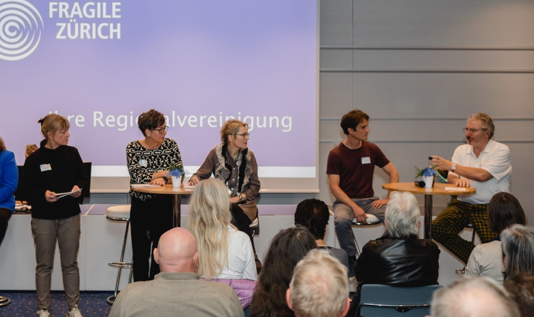 Podiumsdiskussion Ein Foto von hinten aus dem Publikum der Podiumsdiskussion von FRAGILE Zürich. Teilnehmende der Diskussion blicken nach links zu einem Mann im weissen Hemd.